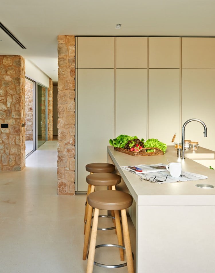 Modern kitchen with beige cabinets and stone accents evokes the feel of luxury travel. A sleek island features wooden stools, a faucet, and is topped with a newspaper, white cup, and fresh greens. Sunlight streams through a large glass door, illuminating the space beautifully. David Pearson Travel