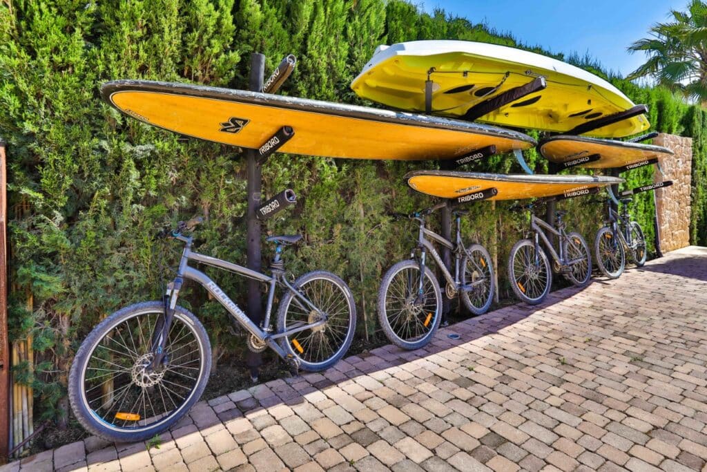 A row of bicycles is parked on a cobblestone path beside a hedge, embodying the charm of luxury travel. Above them, several yellow stand-up paddleboards and a raft are stored on a metal rack. The scene is outdoors, with greenery and a clear blue sky in the background. David Pearson Travel