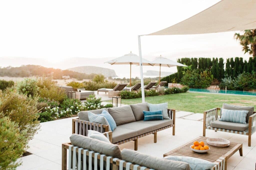 An outdoor patio with cushioned wooden furniture and a coffee table holding a bowl of oranges offers the perfect luxury travel escape. White umbrellas shade lounge chairs by the pool. Enjoy a scenic hillside view with lush greenery and a setting sun as your backdrop. David Pearson Travel