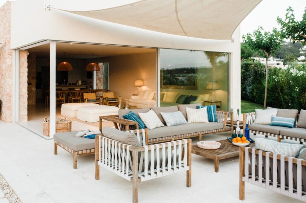 This outdoor patio exudes luxury travel vibes, featuring wooden furniture, white and blue cushions, a beige canopy, and a glass wall leading inside. A table with oranges and a drink complements the lush greenery surrounding this serene retreat. David Pearson Travel