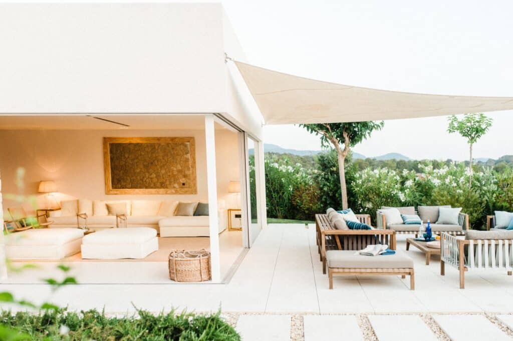 A modern outdoor patio featuring light-colored furniture, including sofas and chairs, offers a taste of luxury travel. The area is shaded by a sail canopy, while an adjacent indoor living room shares the elegant color scheme. Surrounding greenery and small trees add a natural touch. David Pearson Travel