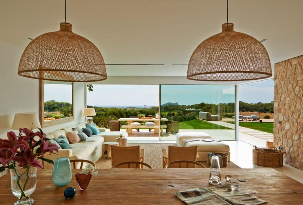 A taste of luxury travel at home, this modern living room boasts large windows with a scenic view. It features a wooden table, wicker chairs, pendant lights, and a cozy seating area with sofa cushions. A vase with flowers and decorative items adorns the table for an inviting atmosphere. David Pearson Travel