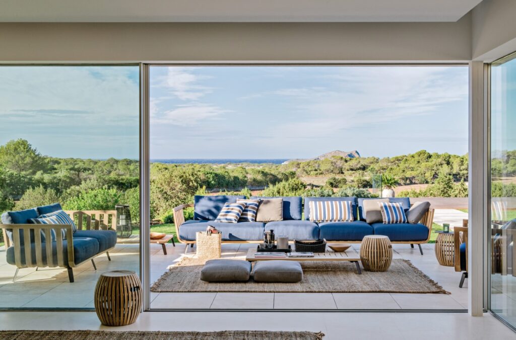 The spacious living room, perfect for luxury travel stays, boasts large glass doors that open to a scenic outdoor view. It features blue and white furniture, including sofas and cushions, natural fiber rugs, and wooden accents. Lush greenery and distant hills are visible outside. David Pearson Travel