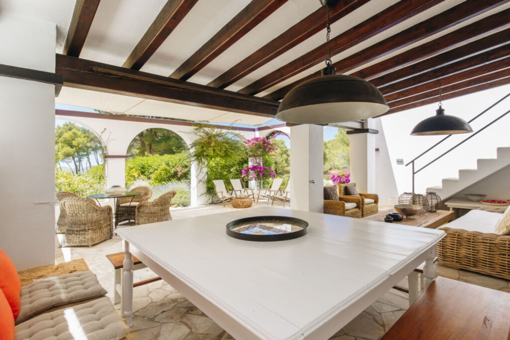 An open-air patio with a large white table under a wooden-beamed ceiling exudes the charm of luxury travel. Wicker chairs and a bench surround the table, while blooming bougainvillea and trees paint the backdrop. Two large pendant lights add an elegant touch overhead. David Pearson Travel