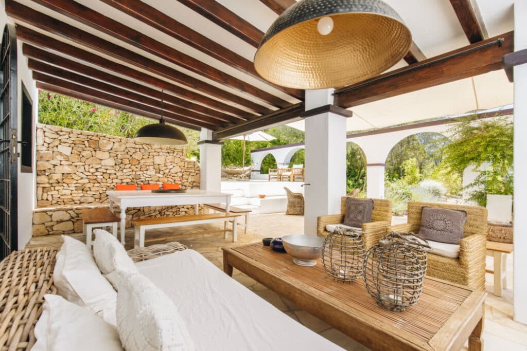 The covered outdoor patio exudes luxury travel vibes with its wooden beams and wicker furniture, including a sofa and chairs. A wooden coffee table displays decorative baskets, while a dining table with a long bench sits in the background. A stone wall and greenery enhance the serene atmosphere. David Pearson Travel