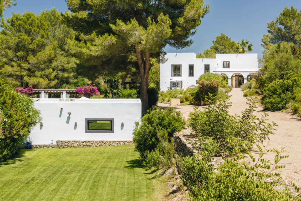 A white stucco house with a flat roof, surrounded by lush greenery and manicured lawns, epitomizes luxury travel beneath a clear blue sky. Bougainvillea flowers adorn the terrace, while a gravel path warmly guides you to the entrance. David Pearson Travel