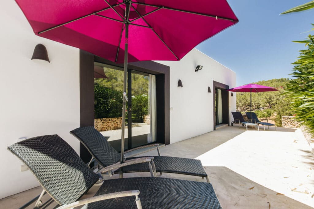 The luxurious patio area features three black wicker lounge chairs and two large purple umbrellas. This elegant seating is set outside a white building with expansive glass doors, surrounded by greenery and a stone wall, all under a clear blue sky—ideal for indulging in luxury travel moments. David Pearson Travel