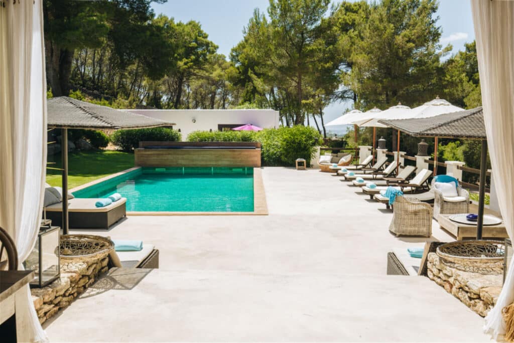 The outdoor swimming pool, surrounded by luxurious sun loungers and umbrellas, is nestled in a lush garden with tall trees. Bathed in sunlight, this idyllic spot embodies the essence of luxury travel, offering a relaxing and serene atmosphere for all who visit. David Pearson Travel