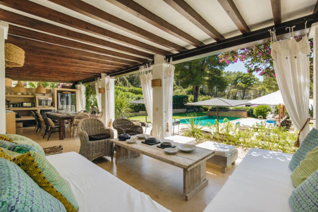Covered outdoor seating area with wicker furniture and a wooden coffee table under a wooden pergola, perfect for luxury travel lounging. Enjoy the view of a lush garden with a swimming pool in the background, while draped white curtains add a cozy, breezy feel. David Pearson Travel