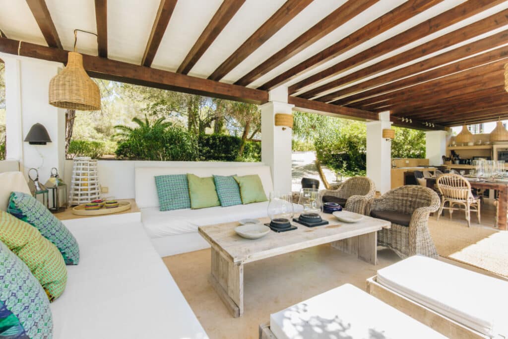 Experience the essence of luxury travel on this outdoor patio, featuring a wooden beamed ceiling and white sofas adorned with colorful cushions. A rustic coffee table and wicker chairs invite relaxation, while an outdoor kitchen enhances the allure. Lush greenery surrounds as sunlight delicately filters through. David Pearson Travel