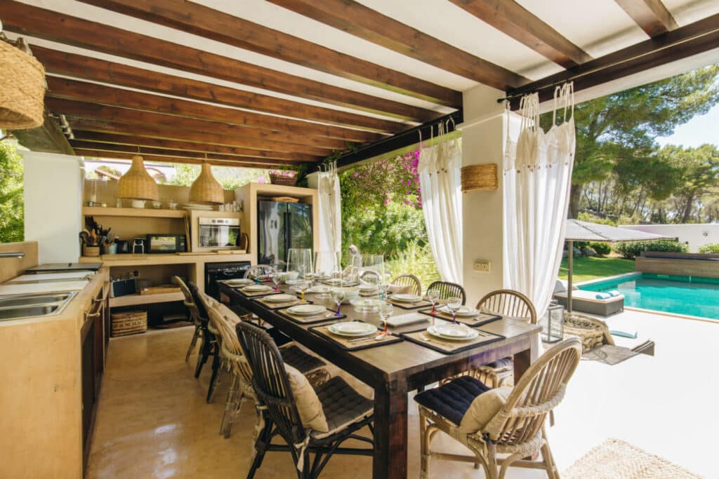 An outdoor dining area exudes luxury travel with its wooden ceiling and wicker chairs around a long table set for a meal. On the left is a modern kitchen, while on the right, an inviting pool is surrounded by lush greenery. Elegant white curtains complete this serene escape. David Pearson Travel