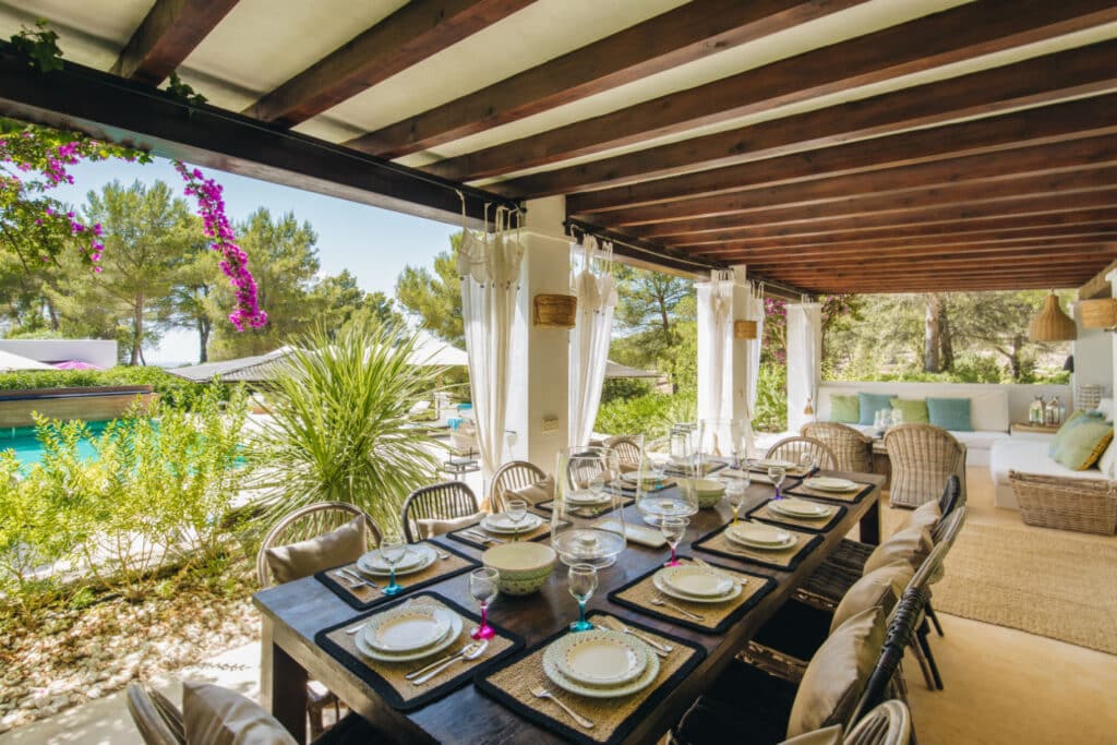 The outdoor dining area exudes luxury travel vibes, with a long wooden table set for a gourmet meal amid elegant plates and glasses. Wicker chairs encircle the setting, nestled under a covered patio embraced by lush greenery. A sparkling pool beckons in the background, framed by purple flowers on the pergola. David Pearson Travel