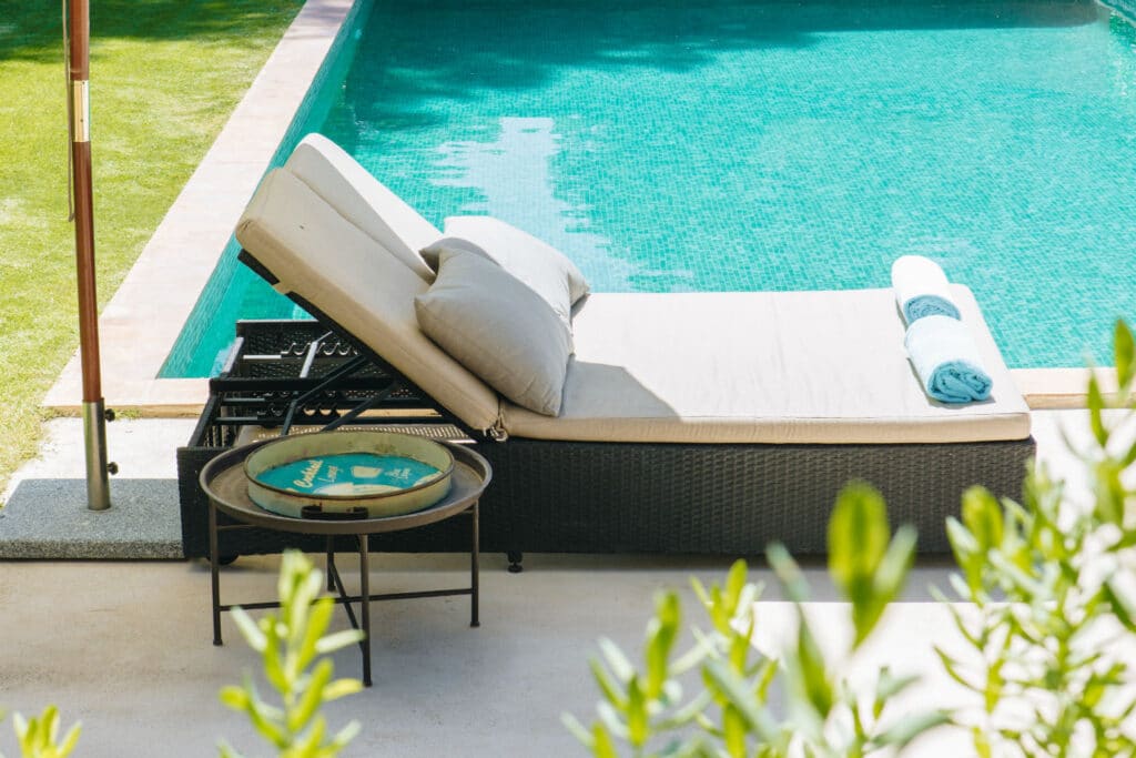A wicker lounge chair with cushions is positioned beside a turquoise pool, epitomizing luxury travel. A neatly rolled towel rests on the chair, while a small table holds a tray with two bottles. Greenery frames the foreground, enhancing this serene setting. David Pearson Travel
