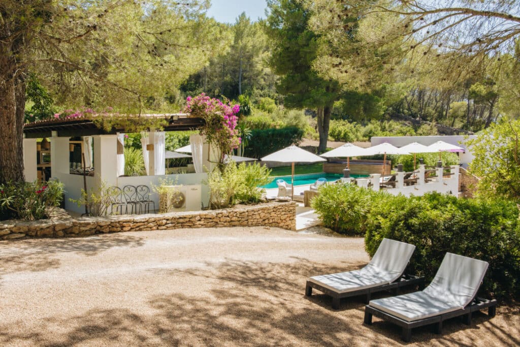A serene outdoor setting ideal for luxury travel features two empty lounge chairs on a gravel path, leading to a stone-walled patio. A canopy-covered seating area and a swimming pool surrounded by umbrellas and greenery promise relaxation, while lush trees frame the background. David Pearson Travel