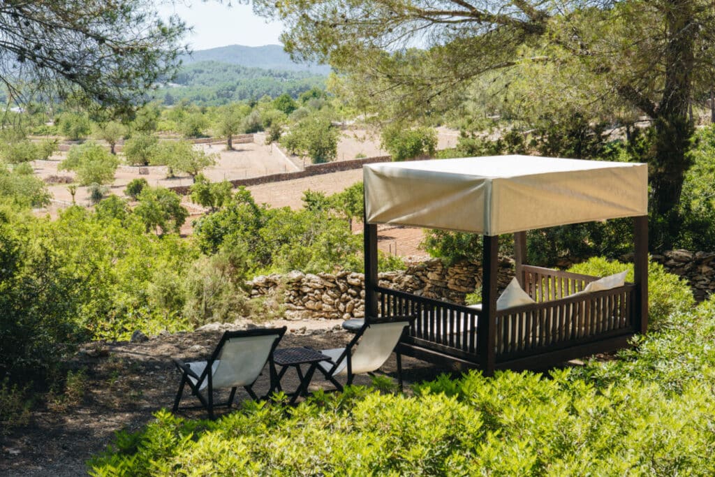 A serene luxury travel experience awaits in an outdoor setting with a cabana featuring plush pillows and two lounge chairs beside a small table. The area, surrounded by lush greenery, overlooks fields and distant hills under a clear sky, offering the perfect escape. David Pearson Travel