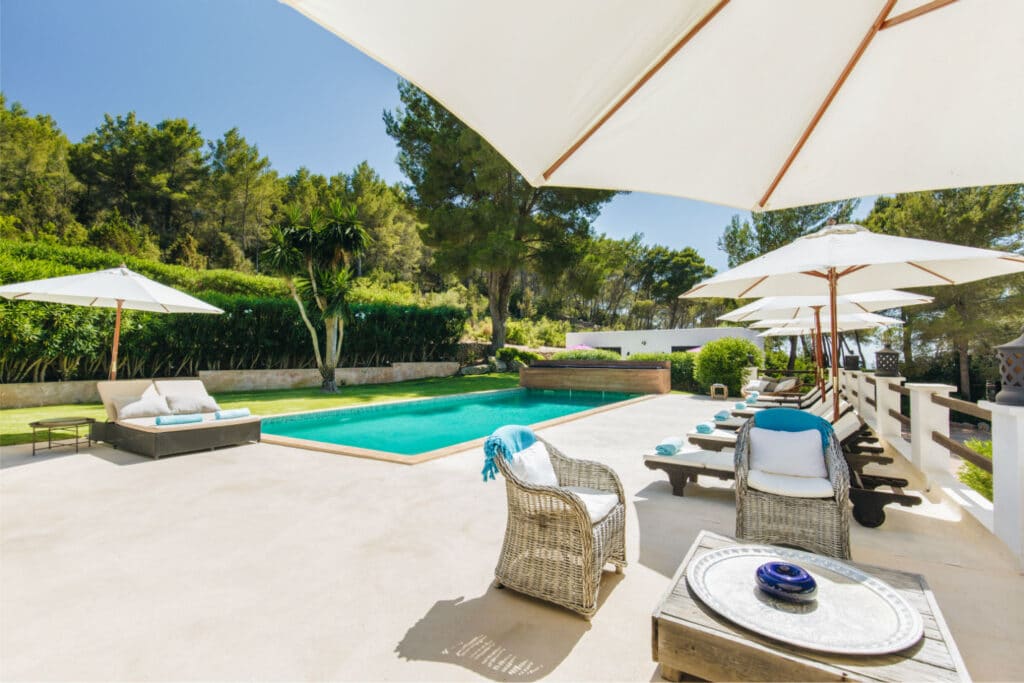 A serene outdoor pool area invites luxury travel enthusiasts with its elegant wicker chairs, sun umbrellas, and lush greenery. The rectangular pool is framed by a light-colored patio, while sunlight filters through tall trees, casting soft shadows that enhance the tranquil ambiance. David Pearson Travel