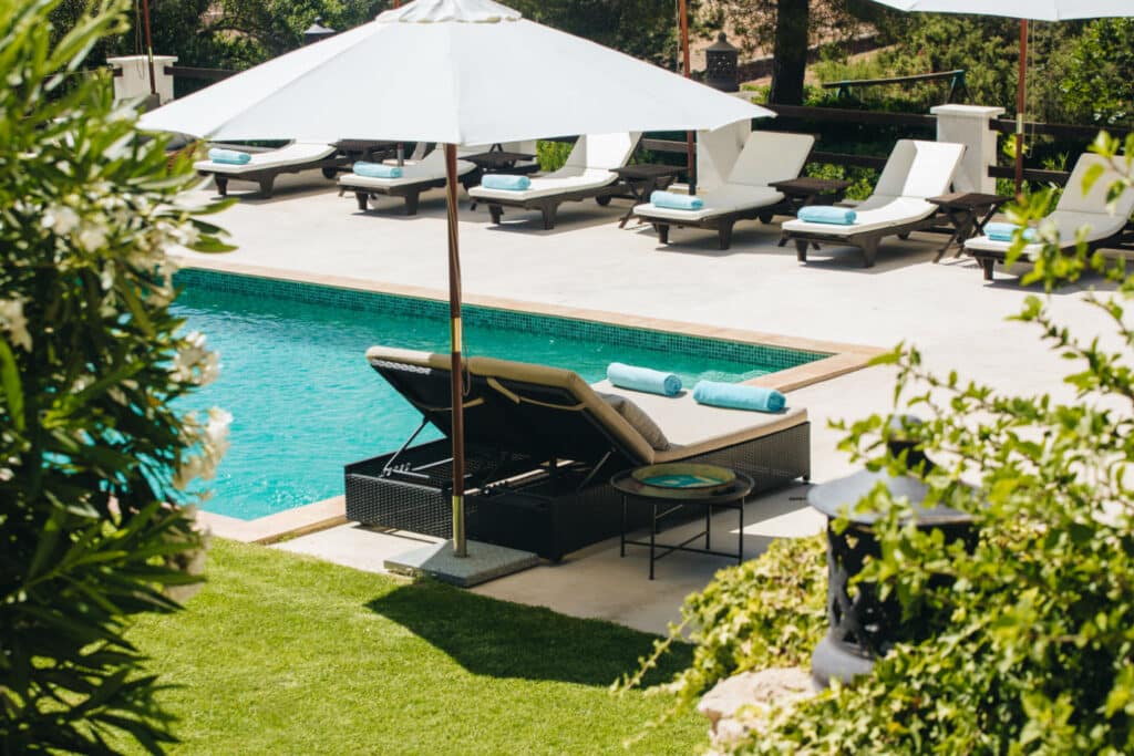 A swimming pool surrounded by loungers with white cushions and blue rolled towels, perfect for a luxury travel experience. Large white umbrellas provide shade, while the pool area is bordered by greenery, creating a serene outdoor setting. David Pearson Travel