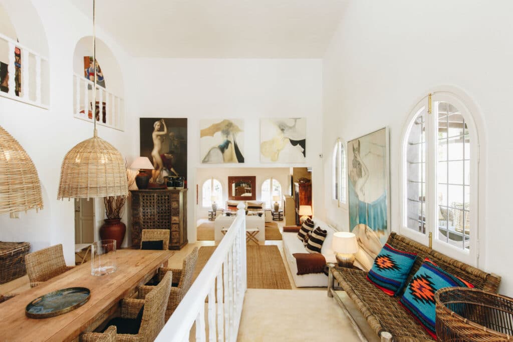 A spacious living room with high ceilings exudes the charm of luxury travel, featuring wicker furniture, a long wooden dining table, and art pieces on pristine white walls. Large arched windows infuse natural light, while woven pendant lights and plush cushions add vibrant touches. David Pearson Travel