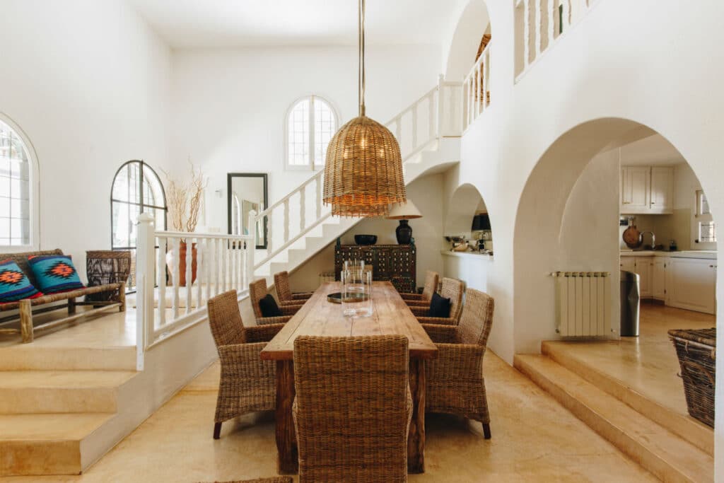 A spacious, bright dining room evokes the elegance of luxury travel with its high ceiling and arched windows. A wooden table is surrounded by wicker chairs, and a large woven pendant light hangs above. Staircase and kitchen are visible in the background, wrapped in warm, neutral tones throughout. David Pearson Travel