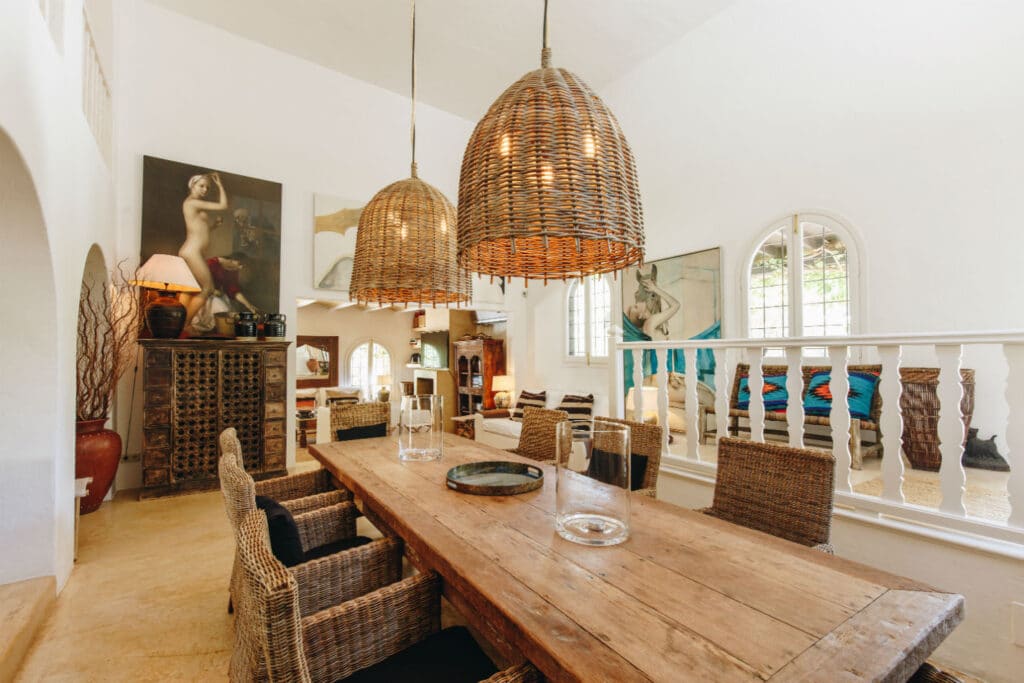 A rustic dining room with a long wooden table surrounded by wicker chairs exudes trendy luxury travel vibes. Two large woven pendant lights hang above, enhancing the ambiance. Art pieces adorn the walls, while an arched window with white shutters lets in natural light, perfect for any elite traveler. David Pearson Travel