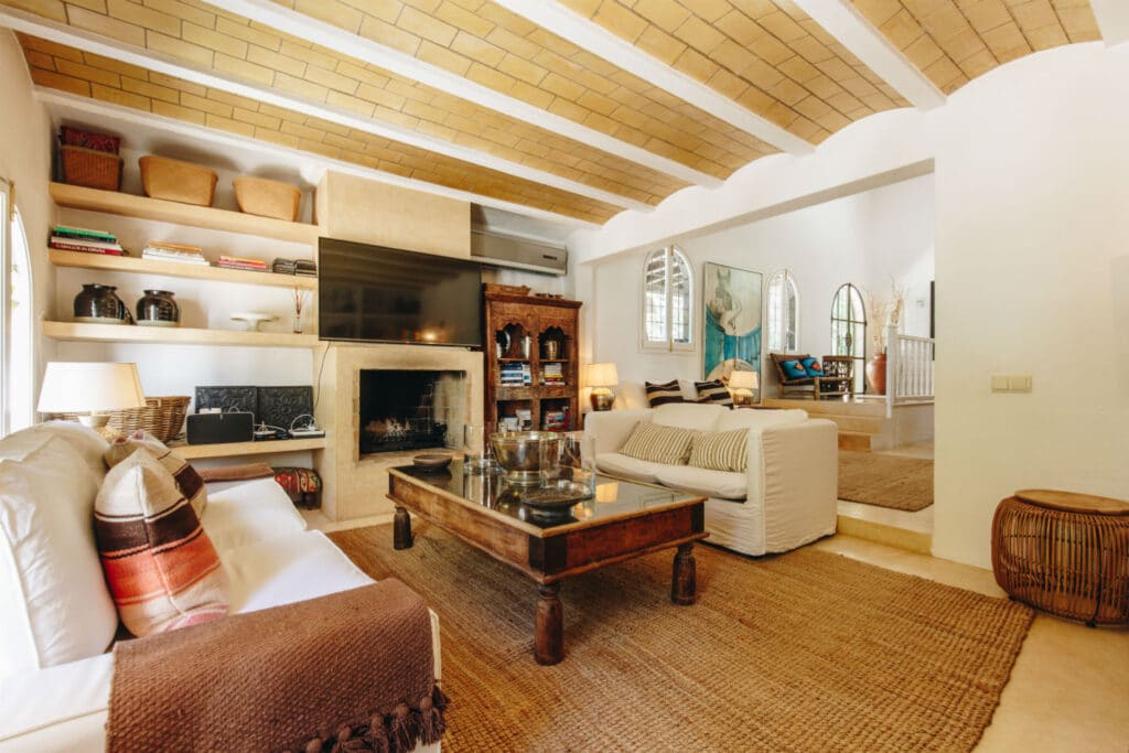 A cozy living room with white sofas, a large wooden coffee table, and a fireplace evokes the indulgence of luxury travel. The walls are adorned with shelves holding books and decor, while the ceiling features wooden beams. A large TV is mounted above the fireplace, and soft lighting creates a warm ambiance. David Pearson Travel