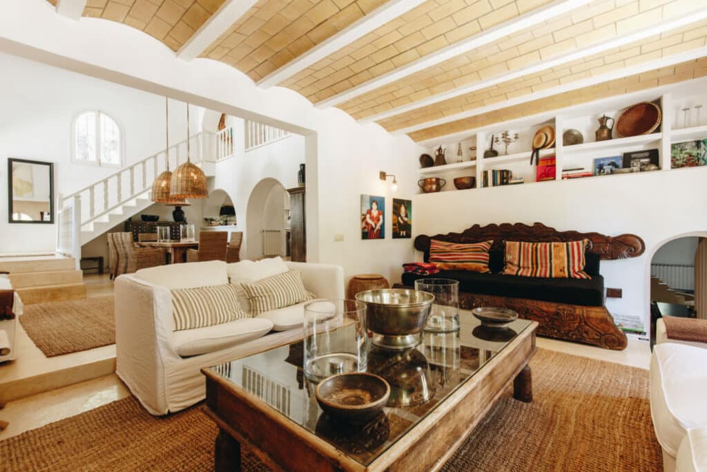 A cozy living room with white walls and a wooden ceiling exudes the charm of luxury travel. It features a plush white sofa, a dark wooden bench adorned with colorful pillows, and a rustic coffee table. Warm lighting enhances decorative items on shelves, set against the backdrop of an elegant staircase. David Pearson Travel