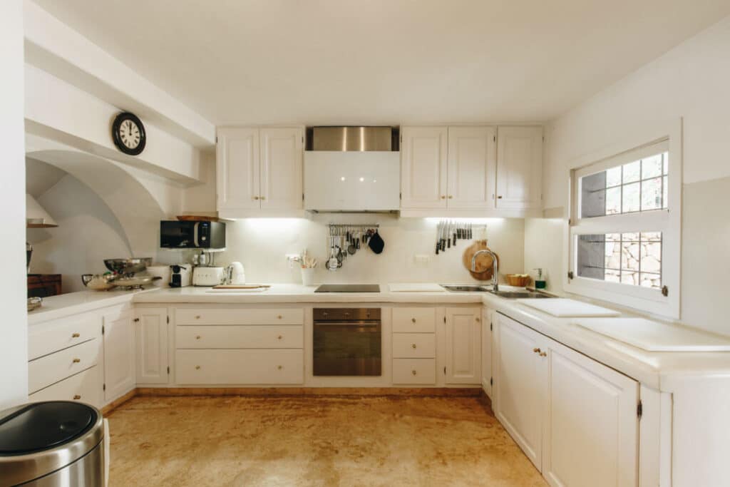 A spacious kitchen with luxury white cabinets, a built-in oven, and a stovetop. Utensils hang above the countertop. A window on the right lets in natural light, while a clock rests elegantly above. A sleek trash can is in the foreground. David Pearson Travel