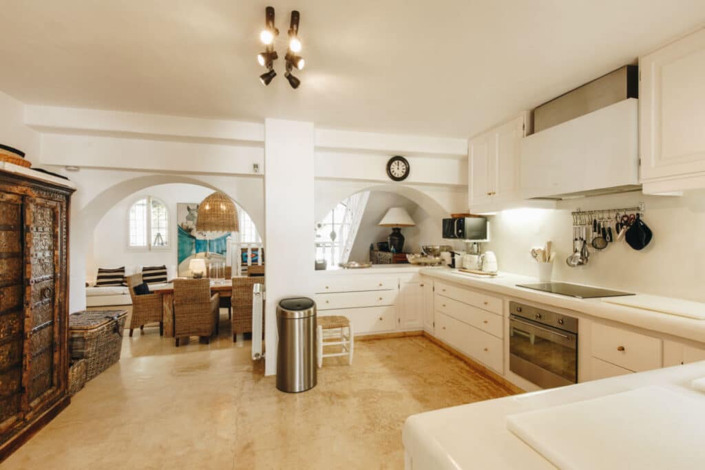 The spacious kitchen features white cabinets and stainless steel appliances, exuding a luxury travel feel with beige flooring reminiscent of serene destinations. Arched doorways lead to a dining area with wicker chairs, a large lamp, and warm touches like a wooden cabinet and wall clock. David Pearson Travel