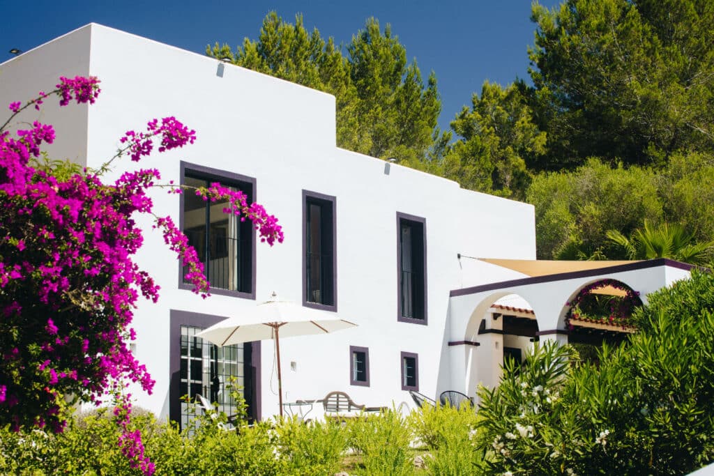 A modern white house with large windows, perfect for luxury travel enthusiasts, is surrounded by lush greenery and vivid purple bougainvillea. There is a patio with chairs, a table, and an umbrella. The background boasts tall trees under a clear blue sky. David Pearson Travel
