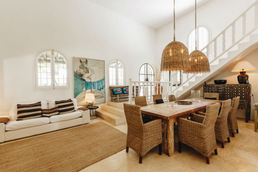 A cozy living and dining area exudes the elegance of luxury travel with wicker chairs around a wooden table under woven pendant lights. A white couch rests on a beige rug, and a staircase with white railings leads up. Arched windows allow natural light to fill the room, enhancing its charm. David Pearson Travel