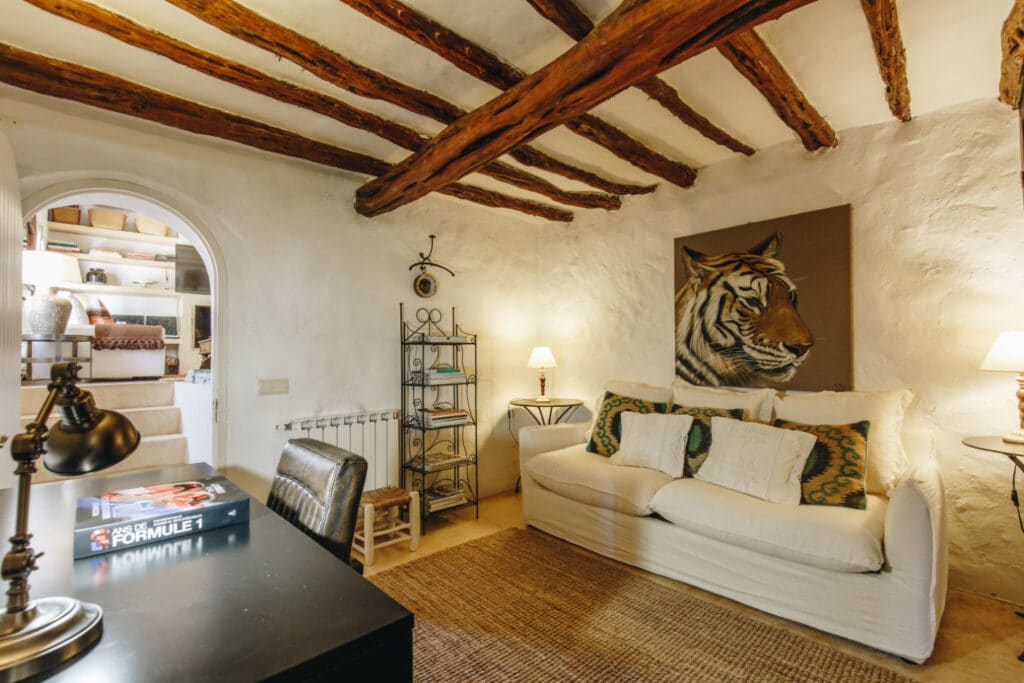 Rustic living room with exposed wooden beams, a white sofa adorned with decorative pillows, and a large tiger painting on the wall. A desk with a lamp and books sits in the foreground, evoking luxury travel vibes. A small arched doorway leads to shelves on the left. David Pearson Travel