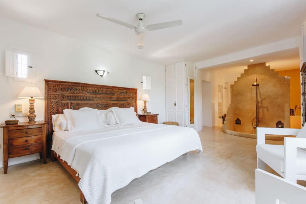 The spacious bedroom, ideal for luxury travel, features a carved wooden headboard on a double bed dressed in white linens. Two matching wooden nightstands with lamps flank the bed. A ceiling fan is overhead, and an open arched entrance leads to a bathroom with a shower. David Pearson Travel