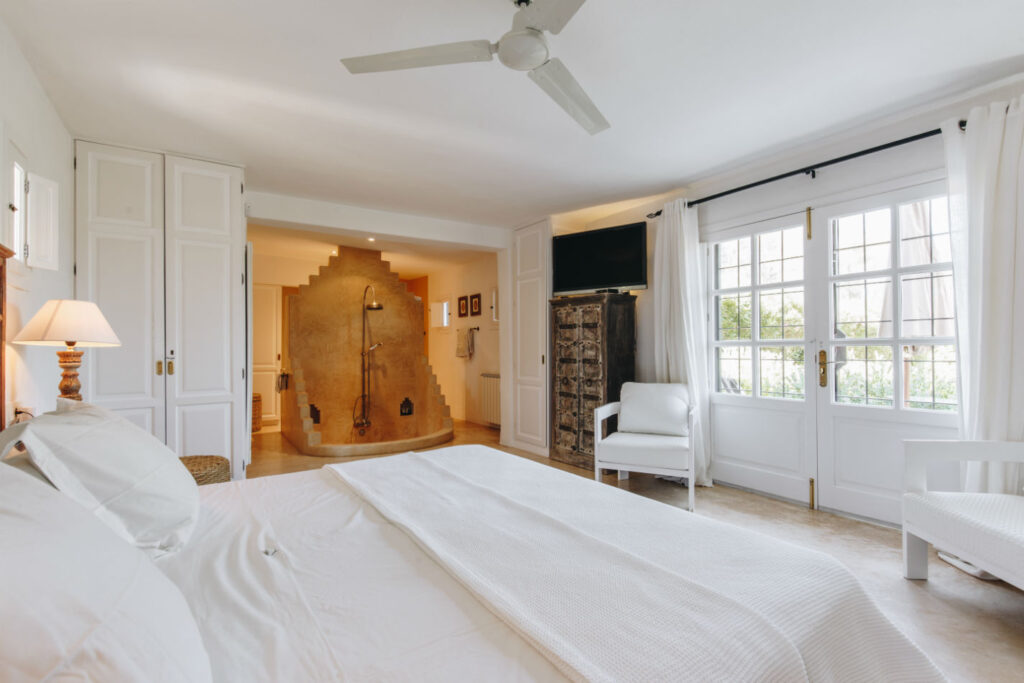A bright, modern bedroom perfect for luxury travel features a large bed with white bedding, a ceiling fan, and a cozy seating area by the window. An open bathroom with a walk-in shower is visible, accented by warm lighting and natural decor. David Pearson Travel