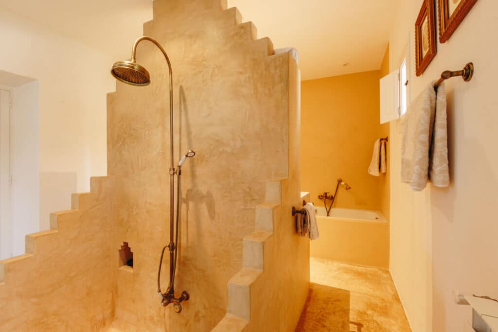 A rustic bathroom exuding luxury travel vibes, featuring a stone-textured shower with a bronze rain showerhead. The walls are a warm beige with stepped edges. In the background, theres an inviting bathtub with plush towels nearby, and picture frames adorn the elegant space. David Pearson Travel