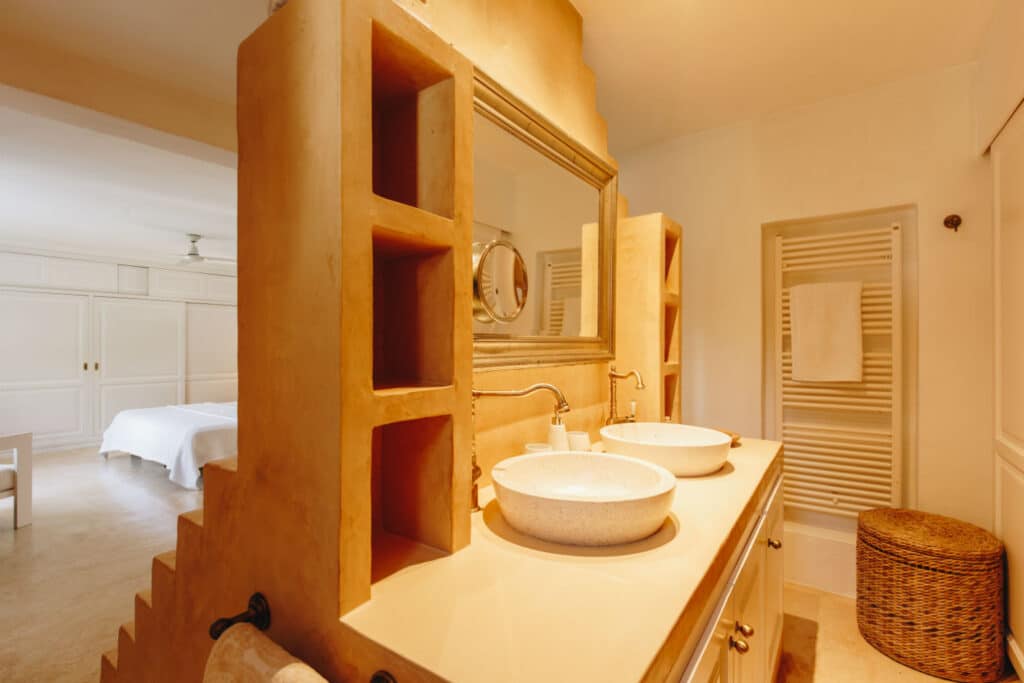 Warm-toned bathroom with double sinks and round mirrors on a wooden vanity. Shelves built into a dividing wall hold small items, adding a touch of luxury travel-inspired elegance. A wicker laundry basket is beside a towel rack, while the bedroom with a white bedspread peeks through in the background. David Pearson Travel