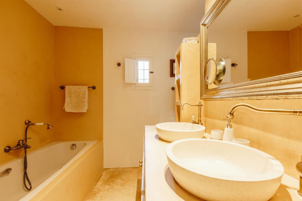 A warm, cozy bathroom exudes luxury travel vibes with a bathtub on the left and a large mirror above a double sink on the right. The light brown walls and soft lighting enhance the relaxing atmosphere, while a towel hangs elegantly on a rack near the bathtub. David Pearson Travel