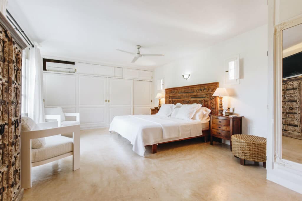 A spacious, bright bedroom perfect for luxury travel, featuring a large bed dressed in white linens. The room includes a ceiling fan, wooden nightstands with lamps, a large mirror, wicker stool, and built-in white closets. Natural light streams through the windows. David Pearson Travel