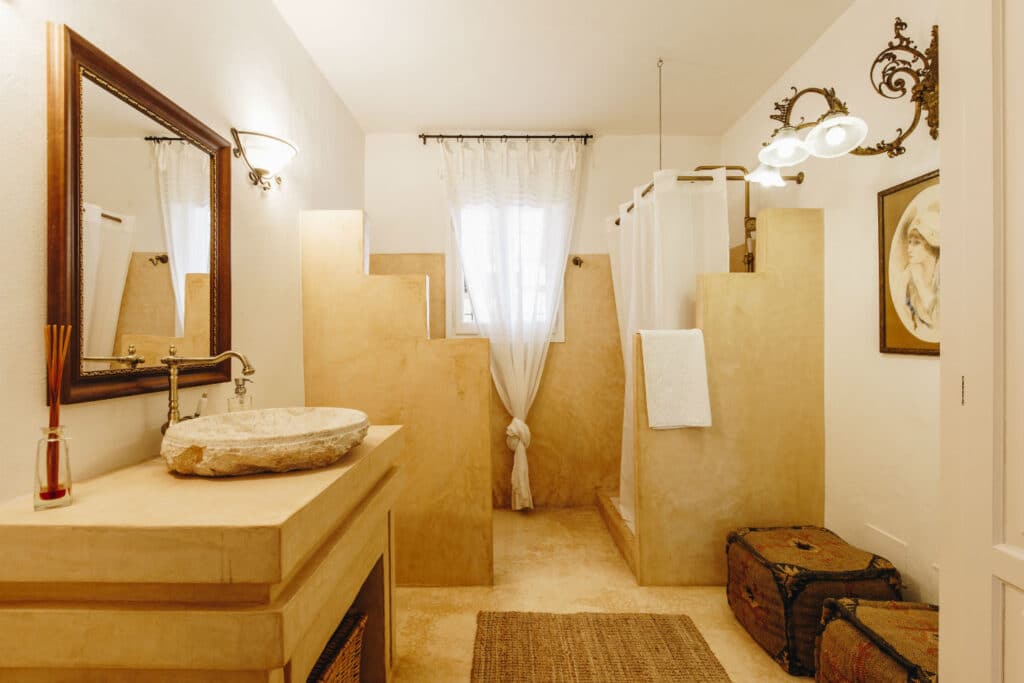 Warm-toned bathroom with a stone sink, decorative lighting, and a unique stepped half-wall shower area evokes the feel of luxury travel. White curtains grace the window, two woven rugs add charm to the floor, and two decorative baskets sit in the corner. David Pearson Travel