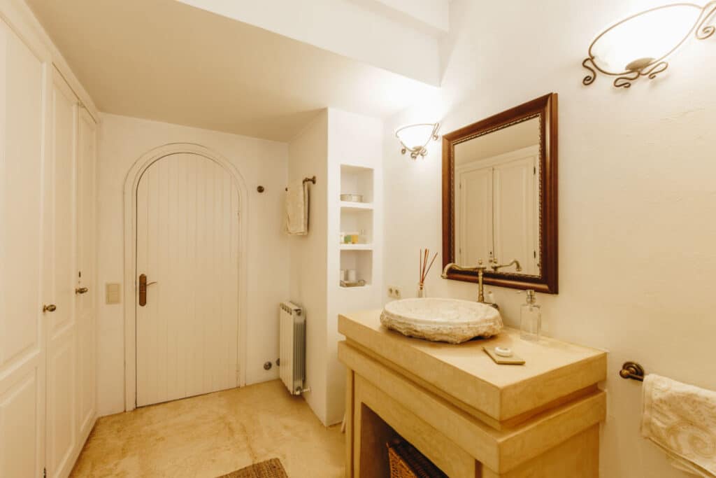 A warm-toned bathroom reminiscent of luxury travel, featuring a rustic sink on a beige vanity. A round mirror hangs above, flanked by elegant wall sconces. A towel graces the nearby wall rack. To the left, a wooden door opens beside small shelves that hold an array of carefully curated items. David Pearson Travel