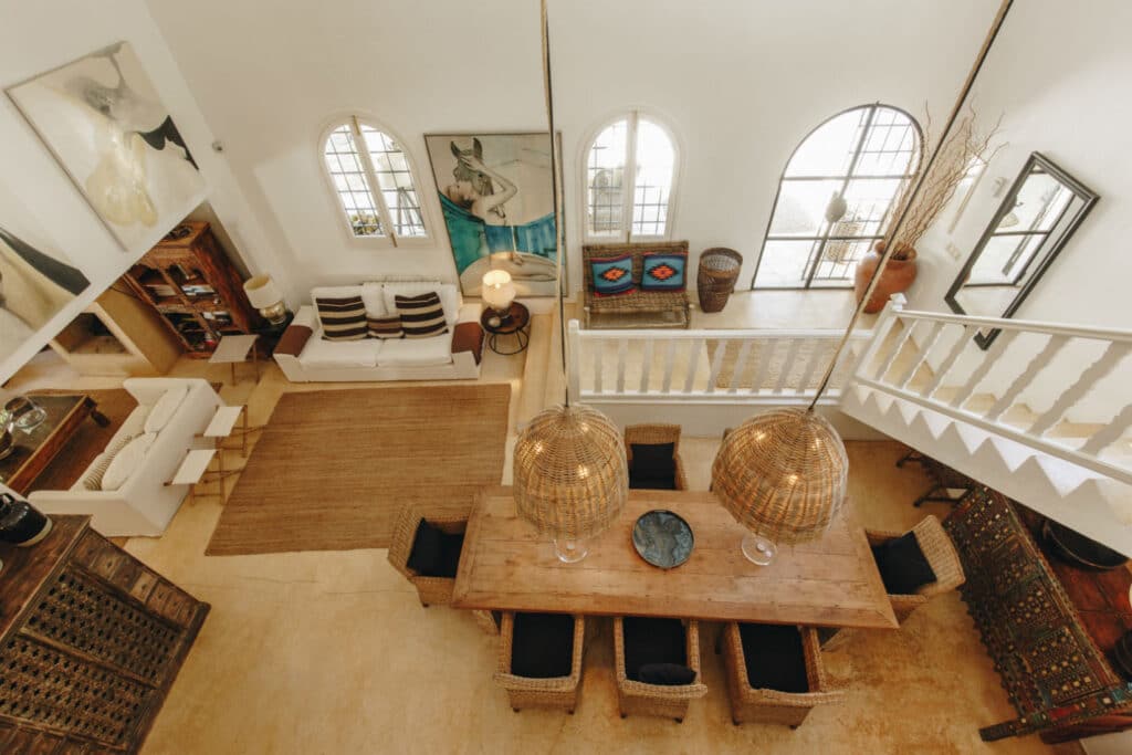 A spacious living room with a high ceiling exudes luxury travel vibes, featuring a wooden dining table, wicker chairs, and two large hanging lights. A white sofa and wooden cabinet sit to the side. Arched windows invite natural light, enhancing the warm, rustic decor. David Pearson Travel