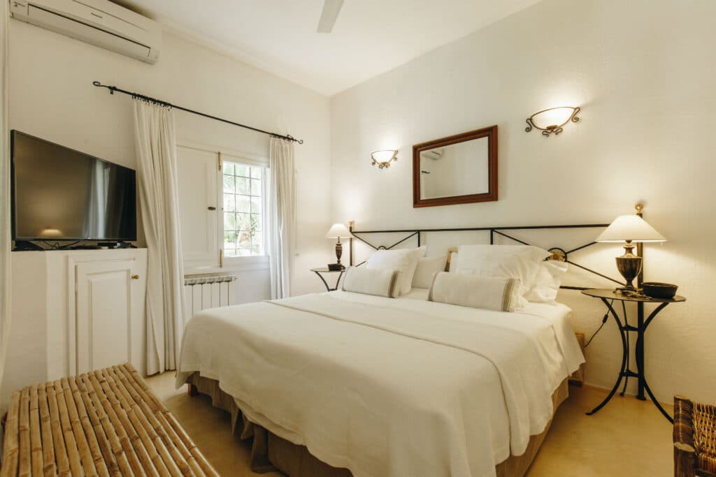 A cozy bedroom reminiscent of luxury travel, featuring a large bed with white linens and numerous pillows. The room includes two elegant bedside tables with lamps, a wall-mounted TV, a window with curtains, and a decorative mirror above the bed. The decor is warm and inviting. David Pearson Travel