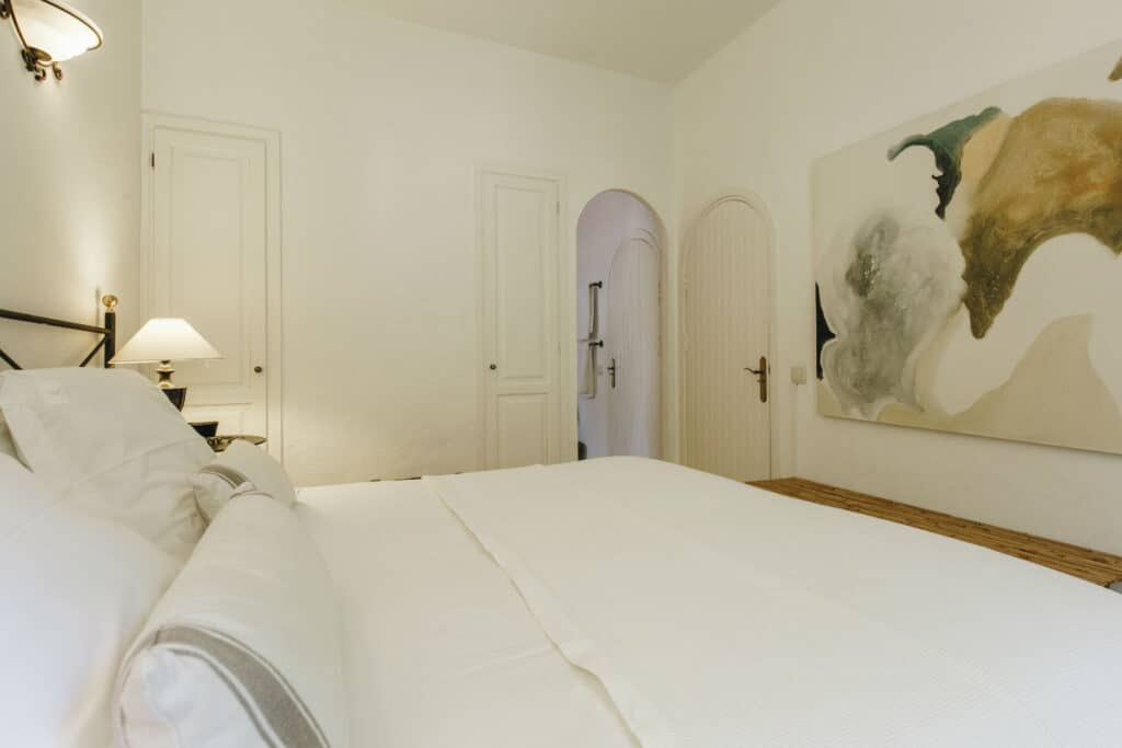 A minimalist bedroom with a large white bed, a bedside lamp, and abstract artwork on the wall evokes the serene elegance often found in luxury travel accommodations. The room features a wooden door and white walls, creating an oasis of simplicity and peace. David Pearson Travel