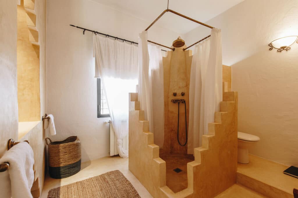 A bathroom with a beige stone step-in shower, surrounded by white curtains, evokes the elegance of luxury travel. It features a woven basket, a textured rug, and a nearby toilet. The space is bathed in soft lighting with a window adorned in sheer curtains. David Pearson Travel