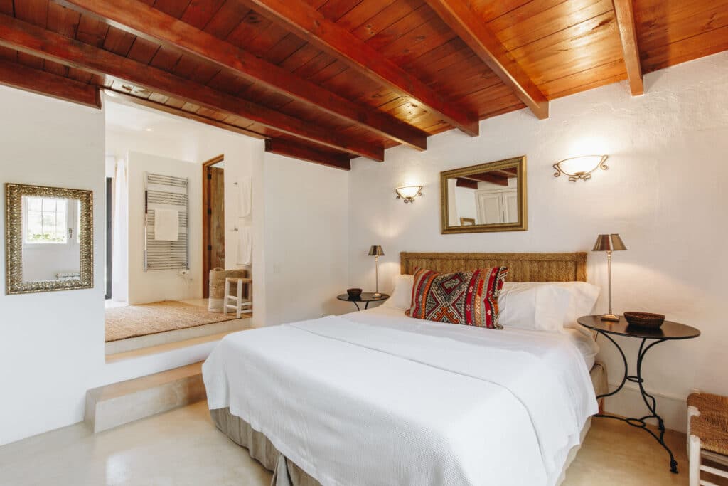 A cozy bedroom with a wooden ceiling, a bed adorned with colorful cushions and a white blanket, exudes the charm of luxury travel. Two side tables with lamps complement wall-mounted lights. A mirror above the bed reflects an open doorway leading to another room featuring an elegant rug. David Pearson Travel