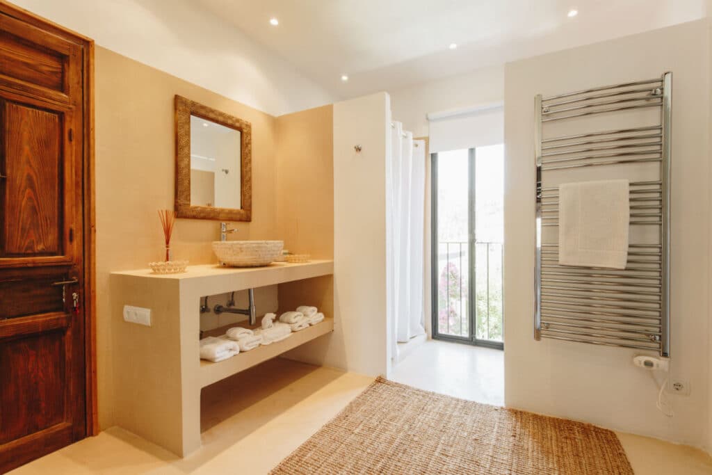 A minimalist bathroom exuding luxury is highlighted by a large wooden-framed mirror above a stone sink. Beneath, shelves display neatly folded towels, evoking a sense of travel serenity. The room features a towel radiator, natural light from the window, and a woven rug gracing the floor. David Pearson Travel