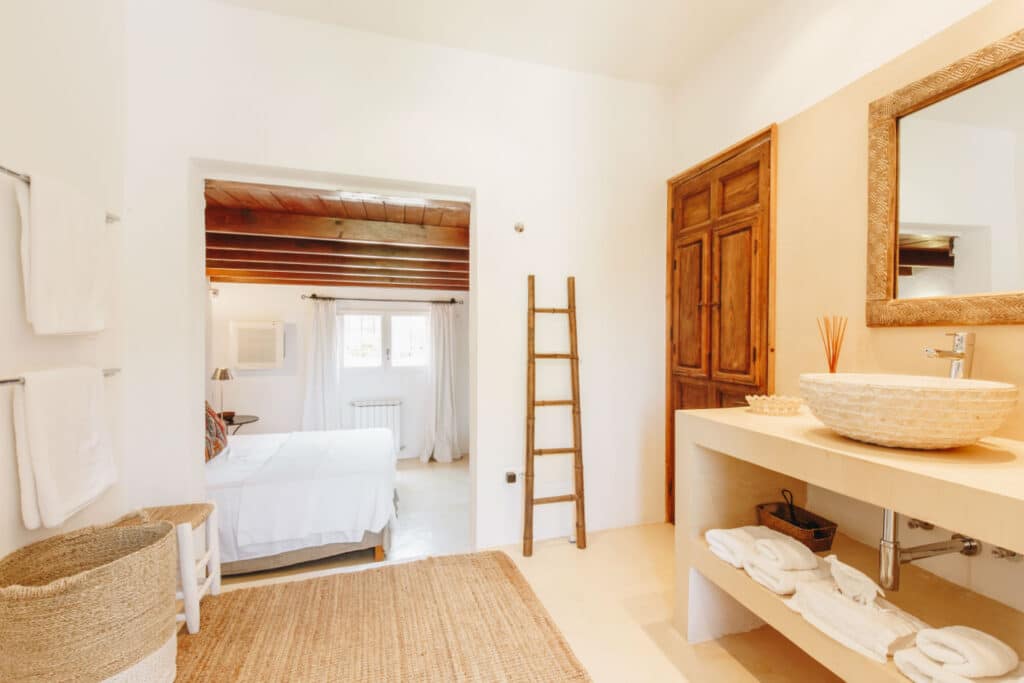 A bright bathroom with white walls exudes luxury travel vibes, featuring a stone sink on a beige countertop, a large mirror, and stylish accents like a wooden ladder and woven basket. Towels are neatly arranged. A doorway opens to a serene bedroom with exposed wooden beams and ample natural light. David Pearson Travel