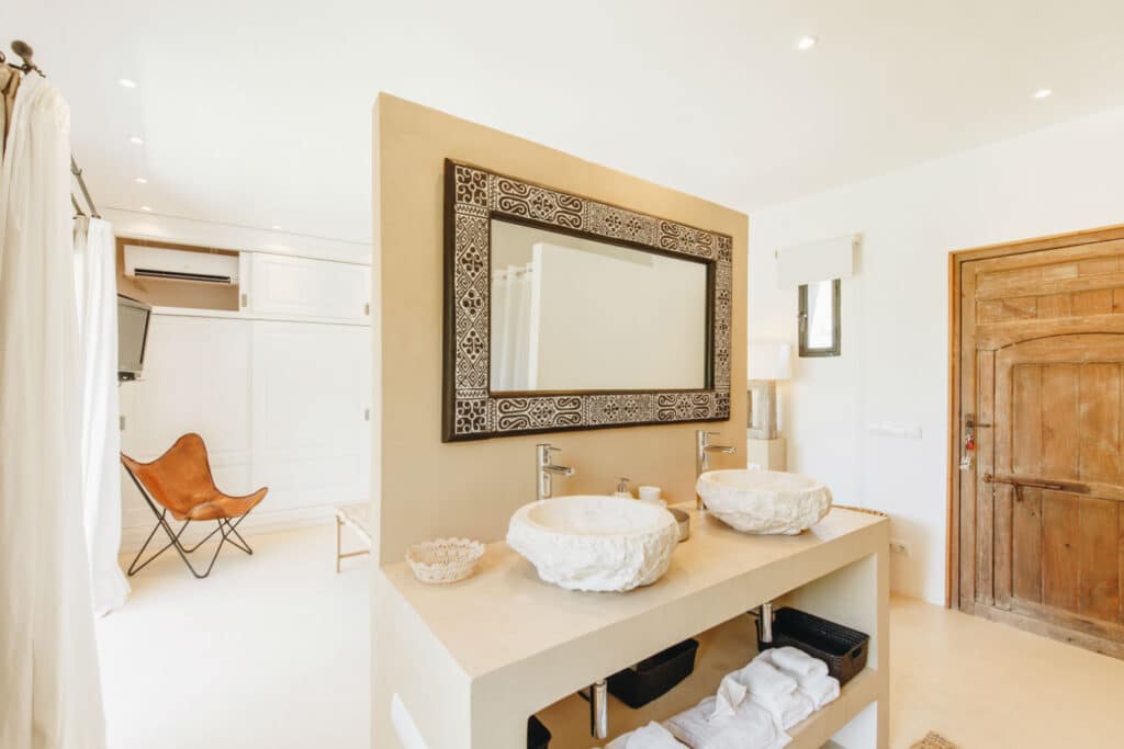 A bright bathroom, perfect for luxury travel enthusiasts, features a large ornate mirror above a double sink vanity with stone basins. Behind the vanity is a wooden folding screen. The room boasts white walls, a wooden door, and a brown butterfly chair in the corner. David Pearson Travel