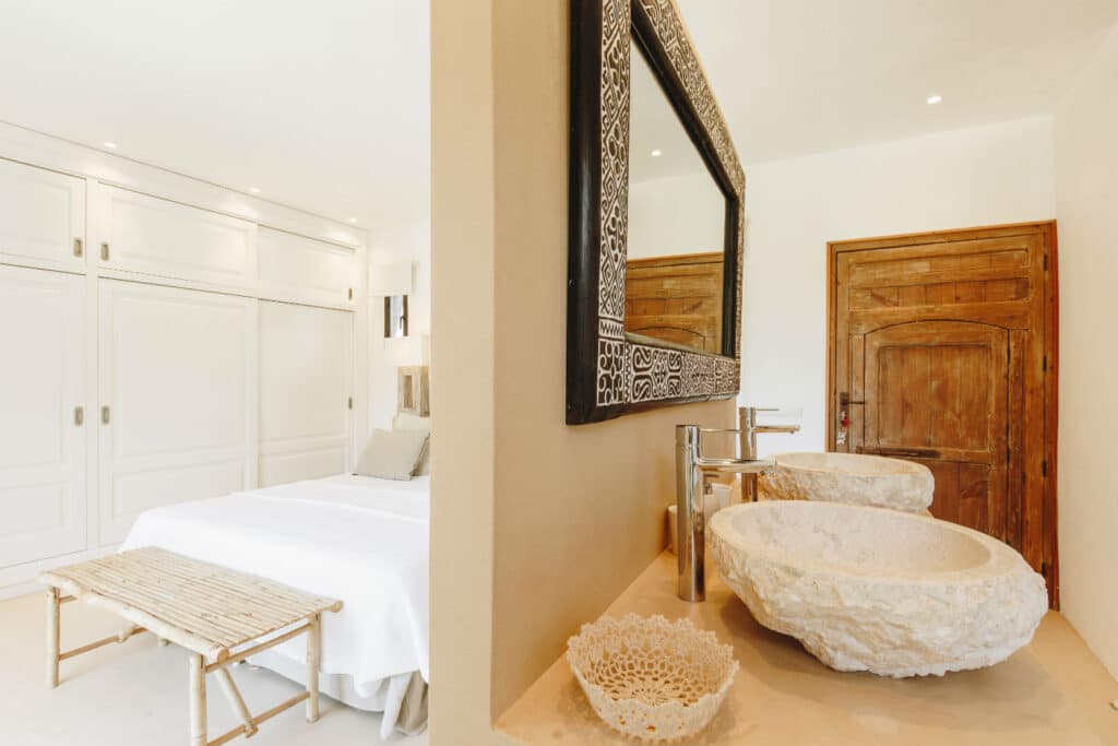 A cozy bedroom with a double bed and white linens is partially visible, adding a touch of luxury travel ambiance. The foreground features a wooden vanity with two stone sinks and a large mirror. A rustic wooden door and white built-in cabinets complete the rooms decor. David Pearson Travel