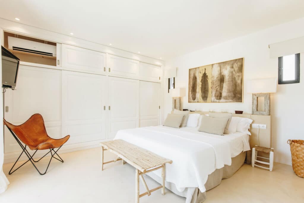 A bright, airy bedroom evokes the charm of luxury travel with its large bed adorned in white linens and beige pillows. A rustic bench graces the foot of the bed, while white built-in cabinets, a wall-mounted TV, a leather chair, and a grand abstract painting add to its elegance. David Pearson Travel