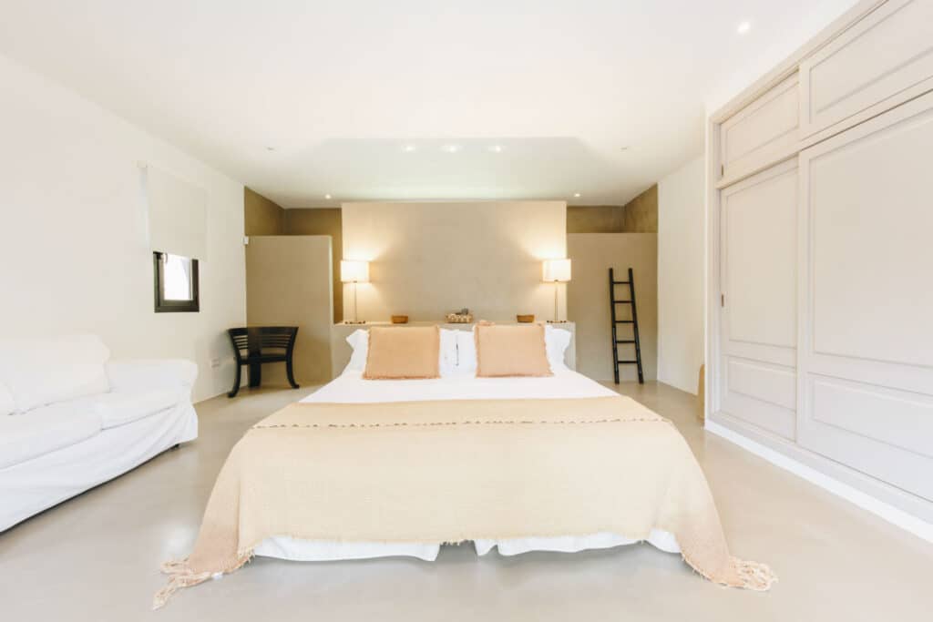 Minimalist bedroom with a large bed featuring beige blankets and pillows, evoking the tranquility of luxury travel. Two lamps flank the bed, with a wooden ladder leaning against the wall. A white couch and black chair are on the left side, and a spacious wardrobe is on the right. David Pearson Travel